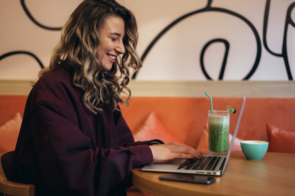 A woman seated at a table, working on a laptop while enjoying a drink beside her.