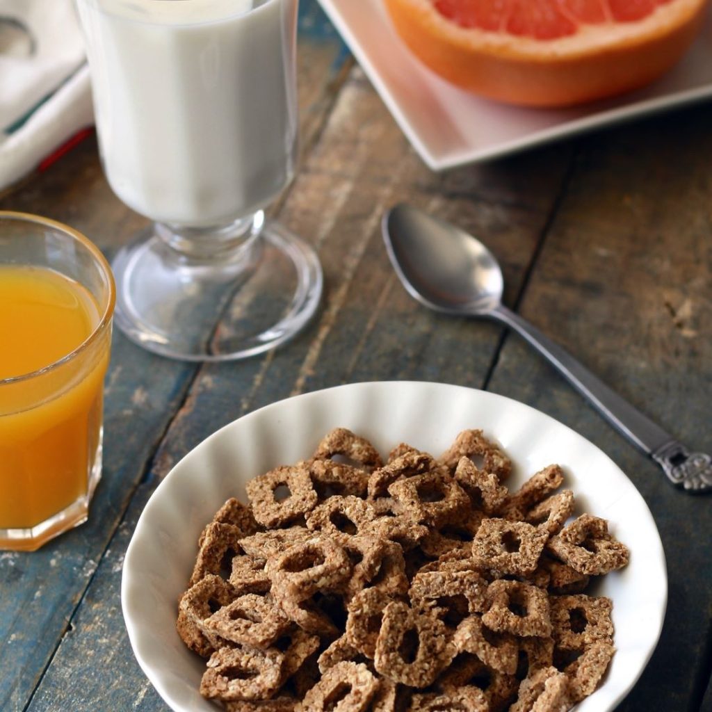A bowl of whole-grain cereal beside a glass of orange juice, a glass of milk, a spoon, and a plate with a grapefruit half on a wooden table.